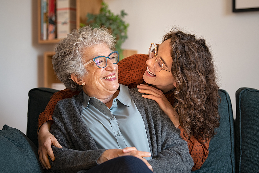 Old grandmother and adult granddaughter hugging at home and looking at each other. Happy senior mother and young daughter embracing with love on sofa. Happy woman hugging from behind grandma with love