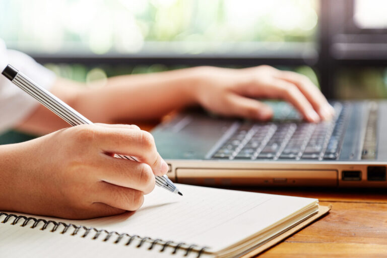 Close up hand of girl writing some data in notebook while studying online course on computer laptop Learning without school or home education concept