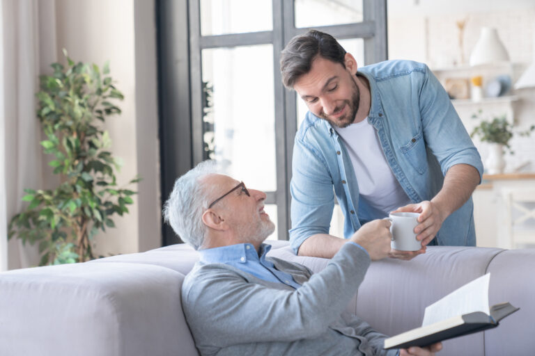 Loving caring young adult son bringing his old elderly senior father a cup of water hot drink at home indoors. Elderly generation support and help aid. Happy father`s day! I love you, dad!